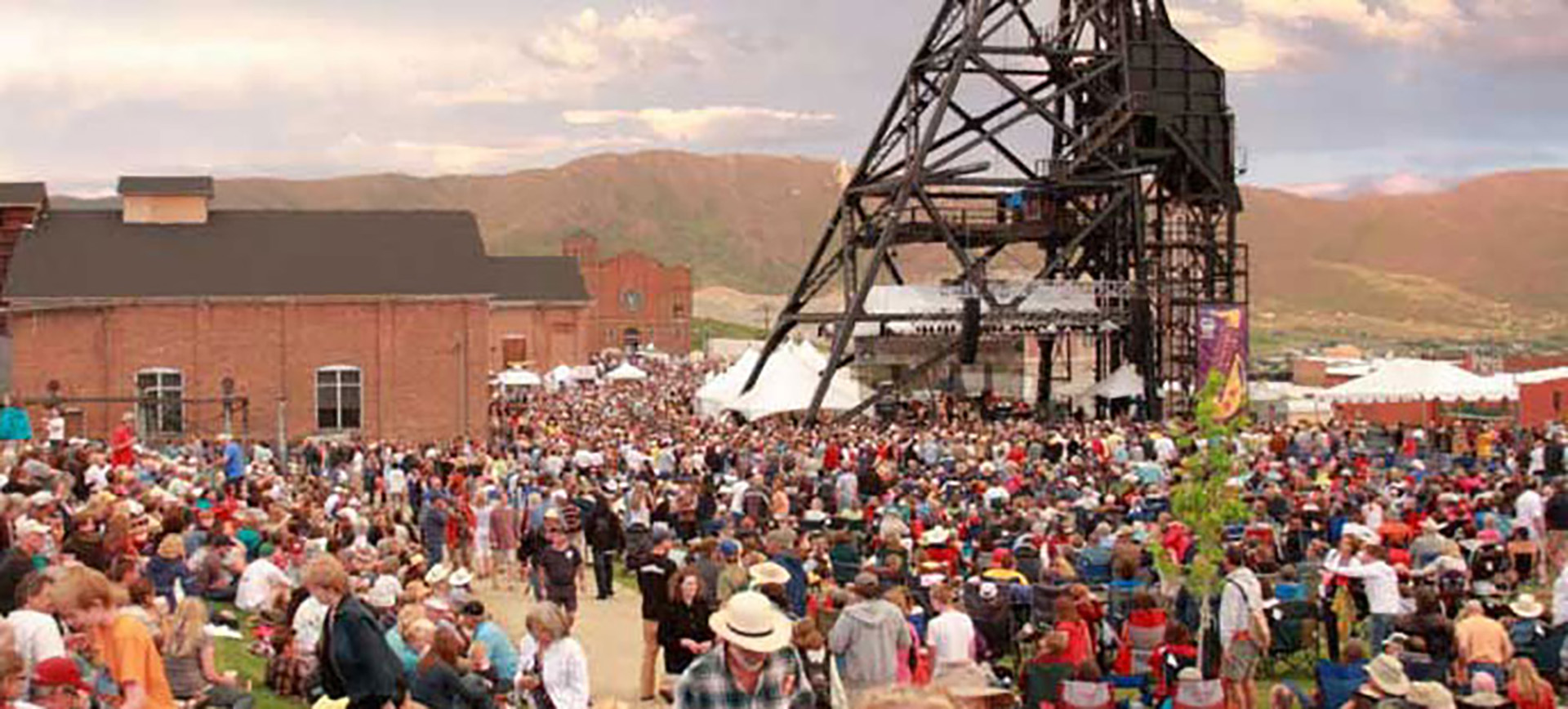 Large outdoor festival crowd gathered beside an industrial headframe and brick buildings with mountains behind